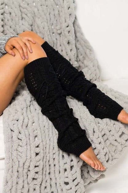 Model wearing Cable Knit Long Tie Leg Warmer in black, sitting on a cozy gray knit blanket.