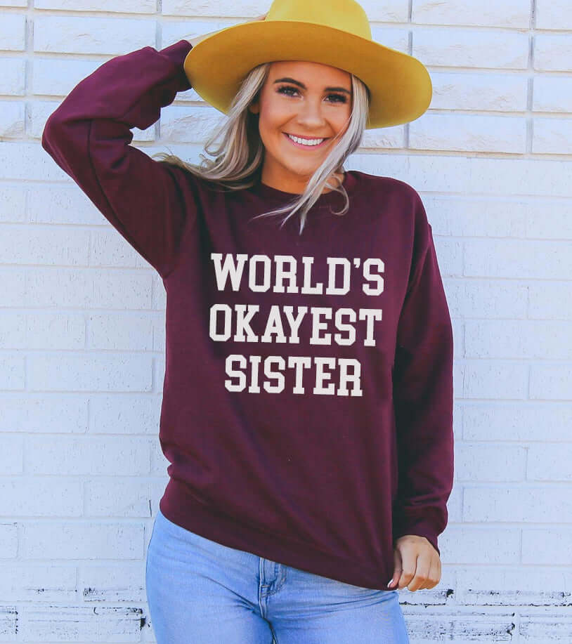 Woman wearing a maroon sweatshirt that reads "WORLD'S OKAYEST SISTER" with a yellow hat and blue jeans.
