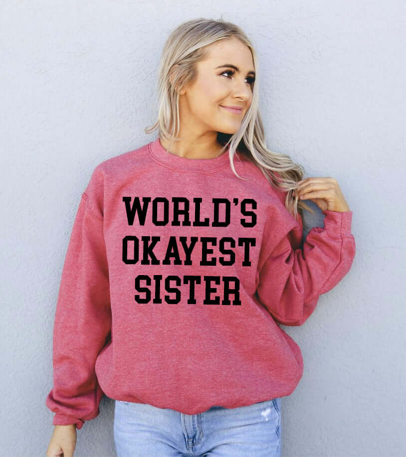 Woman wearing a pink sweatshirt with text 'WORLD'S OKAYEST SISTER' in black letters.