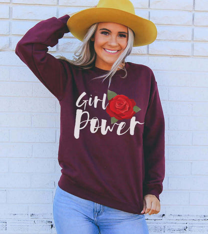 Woman wearing Girl Power Sweatshirt with rose graphic, styled with a yellow hat against a white brick wall.
