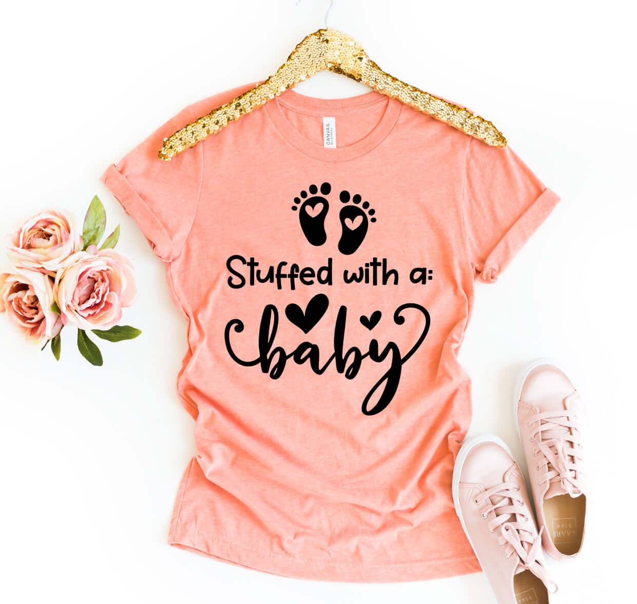 Stuffed With A Baby T-shirt displayed with roses and sneakers, featuring a peach color and playful design.