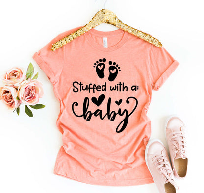 Stuffed With A Baby T-shirt displayed with roses and sneakers, featuring a peach color and playful design.