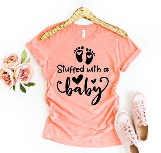 Stuffed With A Baby T-shirt displayed with roses and sneakers, featuring a peach color and playful design.