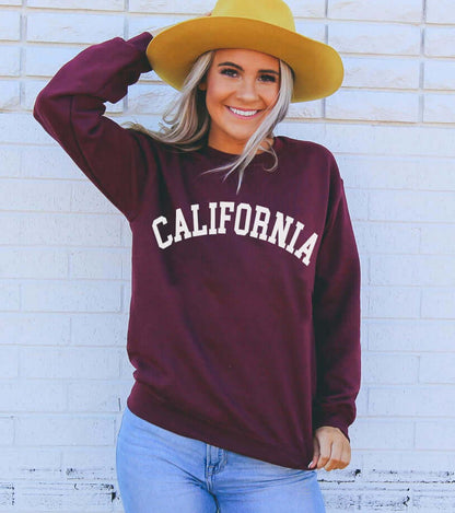 A smiling woman wearing a California Sweatshirt and a yellow hat, posing against a white brick wall.